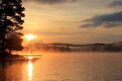Sunrise over Big Moose Lake: mist on the water, a low dock running out from the shore, pines in silhouette against an orange sky