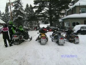 A row of snowmobiles parked in the snow outside the Glenmore in January, riders standing beside them